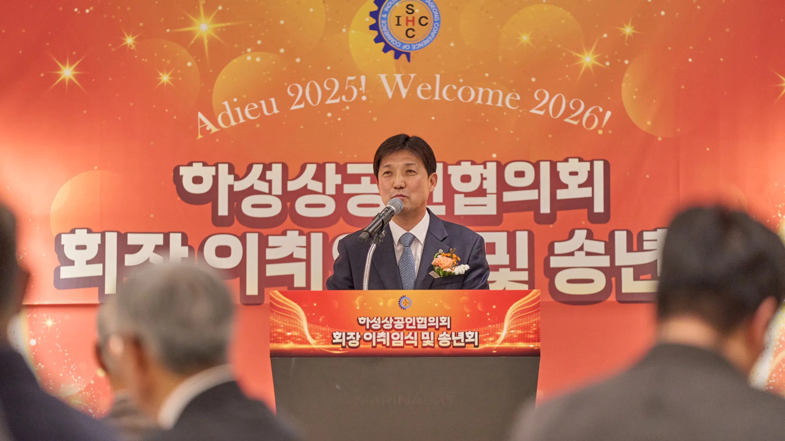 회장 이취임식 무대의 주황색 백월 앞 연단에서 인사말을 하는 회장과 객석의 참석자들