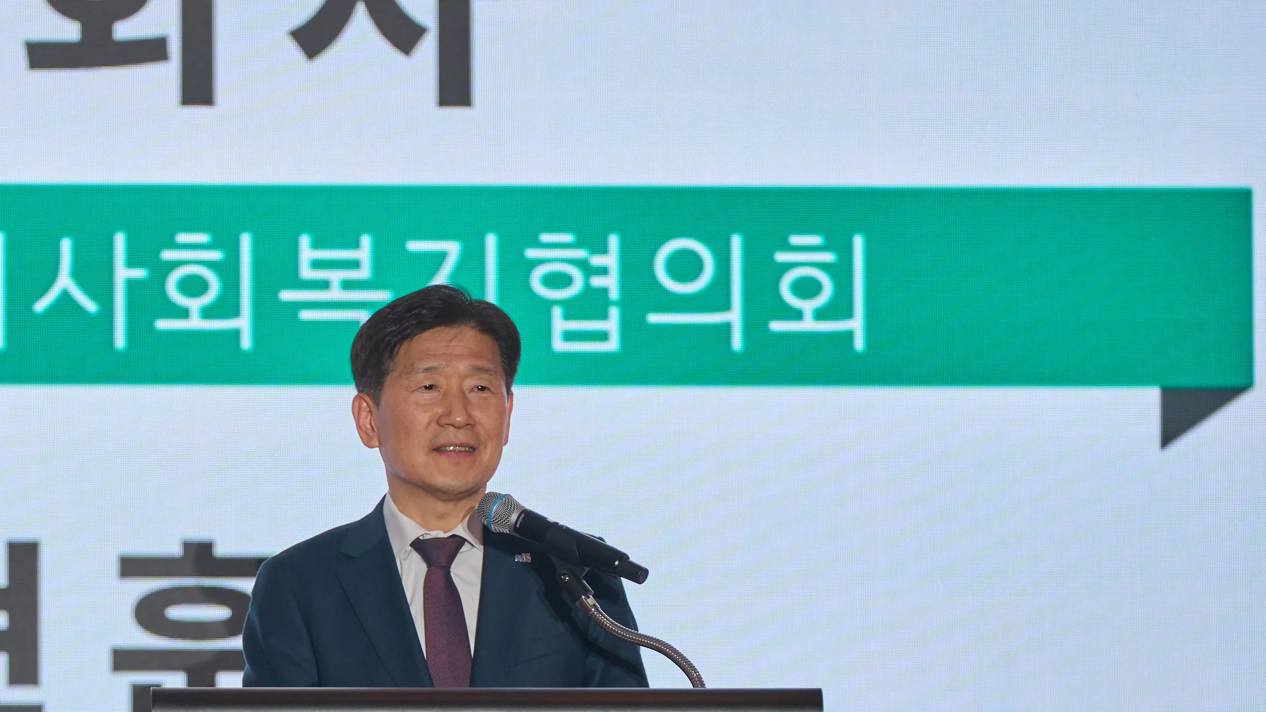 기관 행사 무대의 연단에서 개회사를 하는 연설자와 뒤편 대형 화면의 개회사 자막