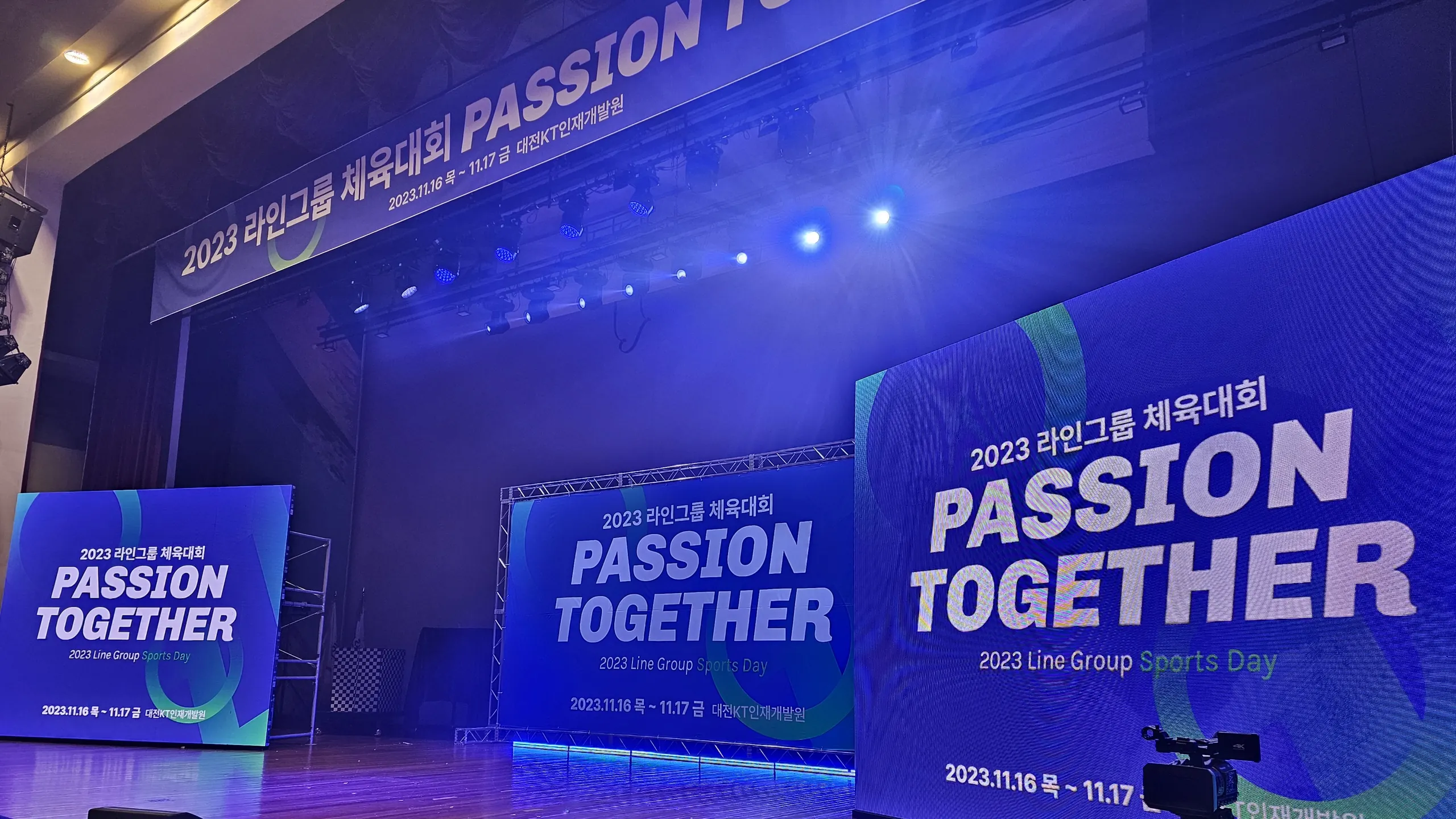 실내 체육관 무대에 체육대회 LED 화면과 배너 세 개가 세워지고 중계 카메라가 놓인 개회 전 모습