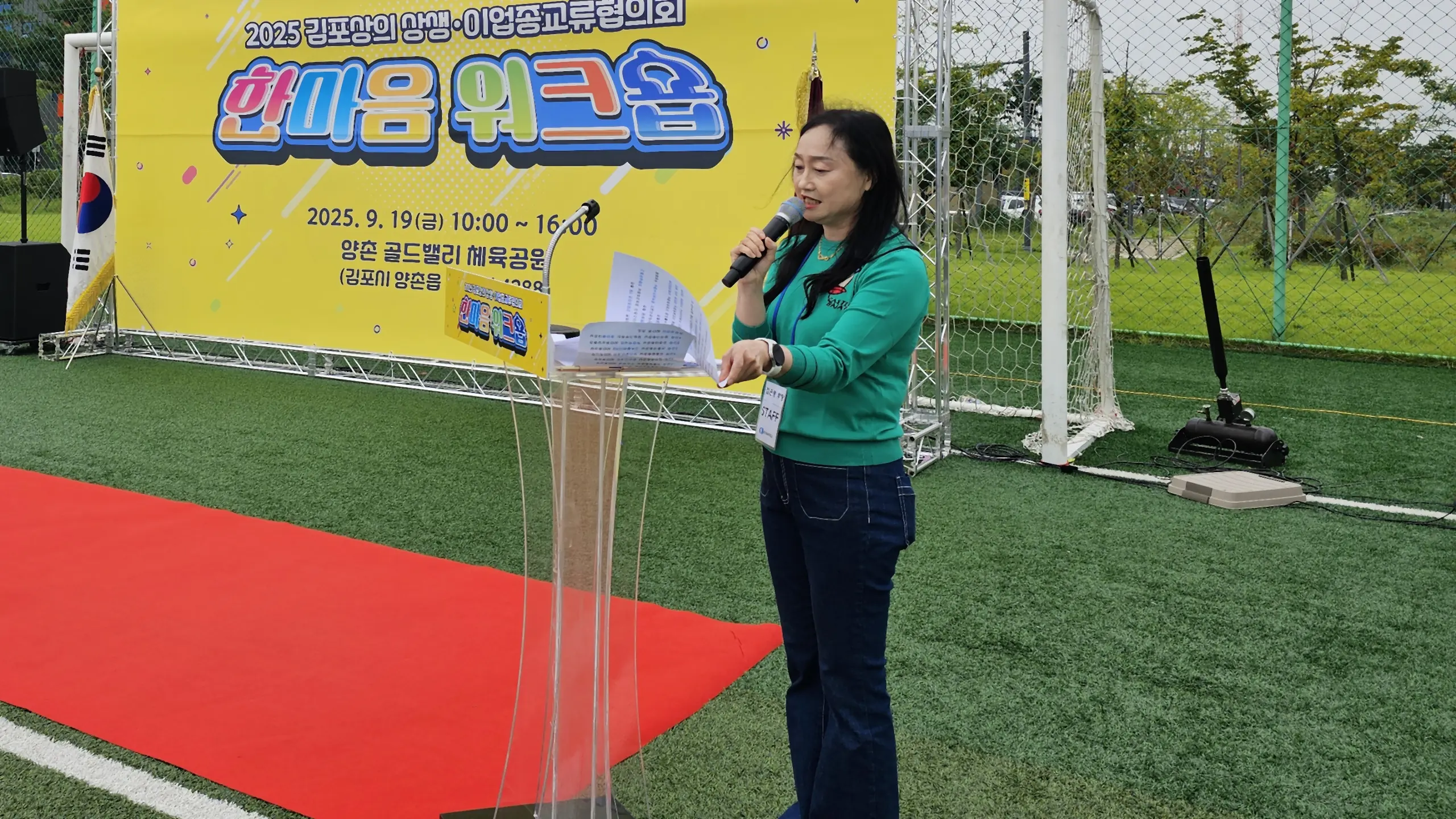 야외 운동장 워크샵 배너 앞 레드카펫에서 무선 마이크로 행사를 진행하는 MC